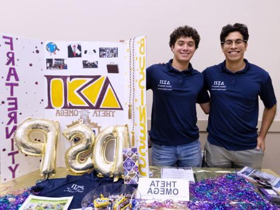 Two students stand behind a table featuring information for their business fraternity at the student involvement fair.